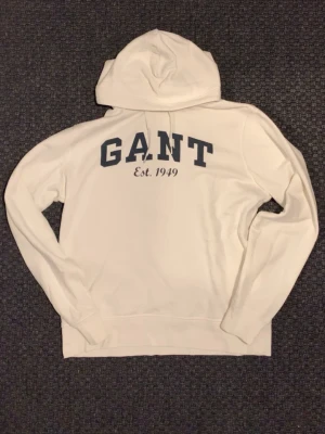 Vit hoodie från GANT med tryck - Säljer en vit hoodie från GANT med stor logga och texten 'Est. 1949' i mörkblått på bröstet. STRLK L men passar även perfekt på M.  Klassisk huva och långärmad modell i mjuk bomull. Perfekt för en chill och stilren look.
