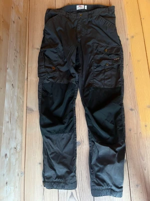 Svarta G1000 friluftsbyxor från Fjällräven - Robusta svarta friluftsbyxor G1000 från Fjällräven med flera praktiska fickor och förstärkta partier på knän och bak. Tillverkade i slitstarkt material, perfekta för vandring och utomhusaktiviteter. Justerbara benslut och klassisk design.