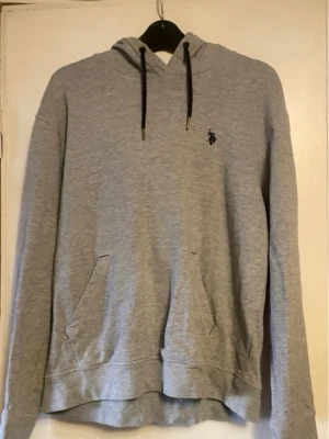 Grå hoodie med ficka och huva - Snygg grå hoodie med svart dragsko i huvan och magficka framtill. Liten broderad logga på bröstet. Tillverkad i mjukt sweatshirtmaterial, perfekt för chill dagar. Långärmad och avslappnad passform.