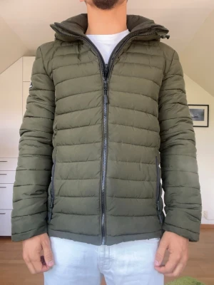 Olivgrön dunjacka från Superdry - Olivgrön dunjacka från Superdry. Luvan går att ta av för en annan look. Strl S. Inga defekter. Modellen är 178 cm och väger 77 kg. Hör gärna av dig vid frågor!