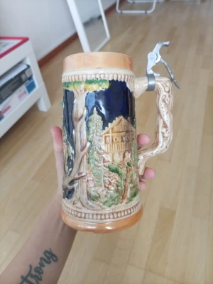 Dekorativ ölsejdel i keramik - Säljer en unik ölsejdel i keramik med handtag och metallock. Sejdeln har ett detaljerat naturmotiv med stuga och träd i grönt, blått och beige. Perfekt för dig som gillar rustik design och samlarprylar. Metallockwt är av tyvärr 