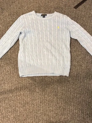Ljusblå kabelstickad tröja Polo Ralph Lauren - Ljusblå kabelstickad tröja från Polo Ralph Lauren med rund halsringning och gul broderad logga på bröstet. 14-16 sitter som xs , Ny skick på tröjan mitt pris 349 kr kom privat vid intresse 