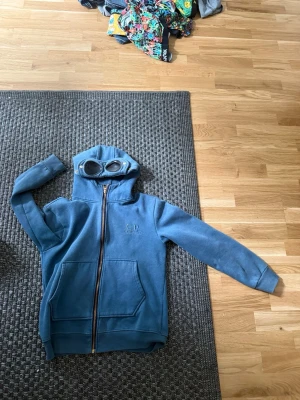 Blå hoodie cp company xs - Säljer en blå zip-hoodie från C.P. Company med ikoniska glasögon i huvan. Hoodien har två stora fickor framtill, ribbade muddar och dragkedja. Perfekt för dig som vill sticka ut med en unik detalj på din hoodie.