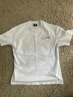 Cortiez Vit t-shirt med tryck - Klassisk vit t-shirt i bomull med rund hals och korta ärmar. På bröstet finns ett diskret tryck i grått. Passar perfekt till jeans eller shorts för en clean och enkel stil.