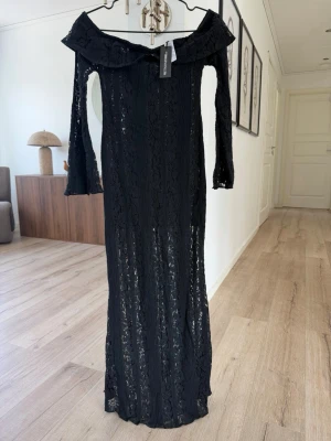 Svart spetsklänning PrettyLittleThing - Lång svart klänning från PrettyLittleThing med offshoulder-design och utsvängda ärmar. Klänningen är täckt av vacker spets och har en figurnära passform som framhäver siluetten. Perfekt för dig som vill ha en elegant och trendig look.