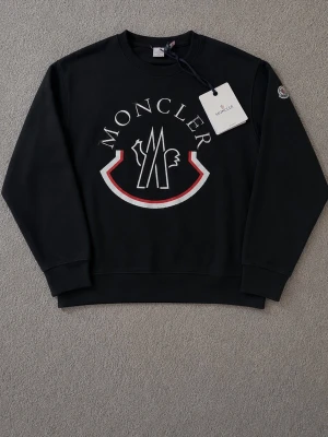 Svart sweatshirt från Moncler - Snygg svart sweatshirt från Moncler med stort vitt och rött logotryck på bröstet. Tröjan har rund hals, långa ärmar och ribbade muddar vid ärmslut och nederkant. Perfekt för en trendig och avslappnad stil.
