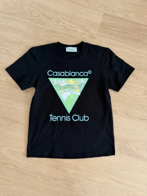 Svart Casablanca Tennis Club t-shirt - Svart t-shirt från Casablanca med tryck på bröstet där det står 'Casablanca Tennis Club' och en färgglad grafisk illustration. Klassisk rund hals och korta ärmar. Perfekt för dig som gillar sportig och trendig stil.