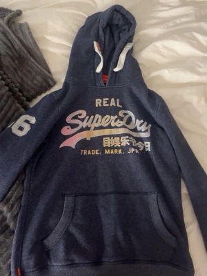 Blå Superdry hoodie med tryck - Villig att sänka priset. Mörkblå hoodie från Superdry med vit och rosa logga framtill, japanska tecken och siffran 6 på ärmen. Klassisk känguruficka och vit dragsko i huvan. Mjuk bomullsblandning och avslappnad passform, perfekt för en chill look.