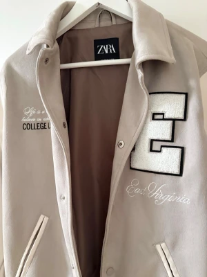 Beige och vit collegejacka Zara - Collegejacka i beige och vitt från Zara med broderad text, stor bokstav E och detaljer som 'East Virginia' och 'College League'. Jackan har ribbade muddar, tryckknappar och två fickor framtill. Materialet är mjukt och jackan har en klassisk varsity-stil.