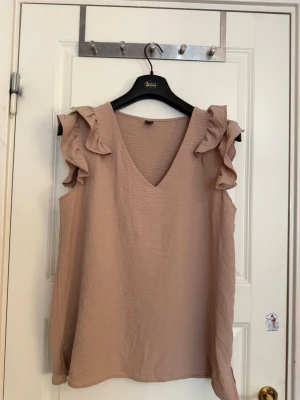 Beige ärmlös blus med volang - Säljer en beige ärmlös blus med v-ringning och volangdetaljer vid axlarna. Toppen har en lös passform och är tillverkad i ett lätt, skrynkligt tyg som känns luftigt och perfekt för varmare dagar.