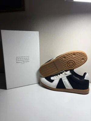 Maison Margiela svartvita sneakers - Maison Margiela sneakers i svart och vitt tyg med vita läderdetaljer och klassisk gummisula i brunt. Lågt skuren modell med vita snören och stilren design som sticker ut. Perfekta för dig som gillar minimalistisk och trendig stil.