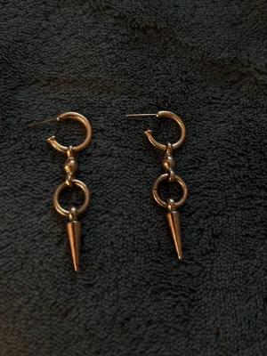 Guldtonade örhängen med konform - Säljer ett par trendiga örhängen i guldtonad metall med rund ring, liten kula och en hängande konformad detalj. Snygg design som ger en edgy vibe till din look. Perfekta för dig som gillar statement-smycken och vill sticka ut.