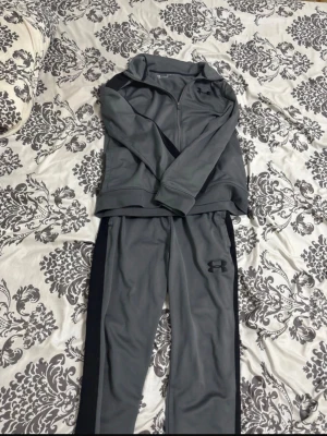 Grå träningsset från Under Armour - Snyggt grått träningsset från Under Armour med ziptröja och matchande byxor. Setet har svarta detaljer längs sidorna på byxorna och en diskret logga på bröstet och benet. Perfekt för gymmet eller löprundan pris kan diskuteras .