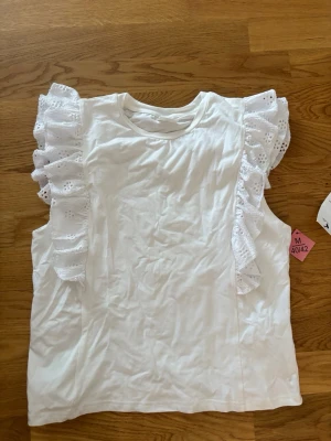 Vit topp med volangärmar - Säljer en söt vit topp med breda volangärmar i broderad spets. Toppen är ärmlös och har rund halsringning. Perfekt för dig som vill ha en feminin och trendig look med extra detaljer på axlarna.