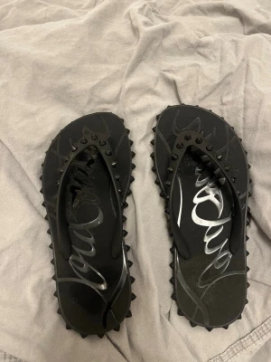 Louboutin sandaler - Säljer ett par svarta sandaler från Louboutin med coola nitar på remmarna och den ikoniska röda sulan. Sandalerna har en chunky look och är tillverkade i gummi med grafiskt mönster på fotbädden. Orginalpris 4000kr med frakt