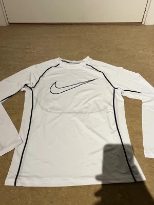 Vit långärmad träningströja Nike - Säljer en vit långärmad träningströja från Nike med svarta detaljer och stor Nike-logga på bröstet. Tröjan har svarta sömmar längs ärmar och sidor, rund hals och är tillverkad i ett stretchigt funktionsmaterial som passar perfekt till träning.