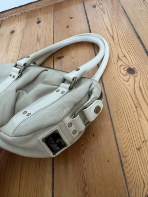 Vit handväska från Tiger of Sweden - Snygg vit/beige  handväska från Tiger of Sweden i mjukt läder. Väskan har dubbla handtag, silverfärgade metalldetaljer och en dekorativ logotypplatta på sidan. Stängs med dragkedja och har en rundad form som ger en modern look. Lite sliten i själva lädret samt lite torr i handtagen, men funkar utmärkt.