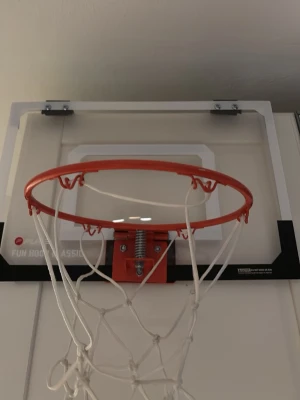 Mini basketkorg för dörr - Säljer en mini basketkorg som enkelt kan hängas på en dörr. Korgen har en orange ring, vit nätkorg och en transparent bakplatta med svart kant. Perfekt för att skjuta lite hoops hemma eller på kontoret.