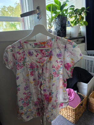 Blommig blus - Söt blommig blus från Flash Woman i vitt med färgglada blommor i rosa, lila, gul och grön. Blusen har korta puffärmar, rund halsringning med knytband och dekorativa veck framtill. Perfekt för en somrig och fräsch look. Hade en reva vid köp som är igensytt, syns knappt 