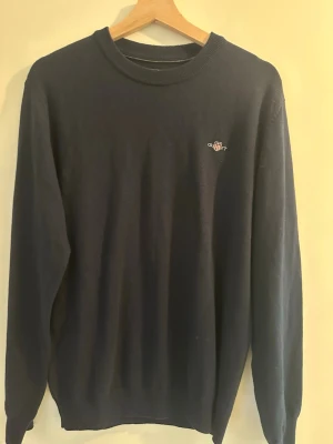 Mörkblå GANT crewneck - Mörkblå GANT crewneck med rund hals och diskret broderad logga på bröstet. Tröjan har långa ärmar och ribbade muddar vid hals, ärmslut och nederkant. Perfekt för lager på lager och enkel att matcha med olika stilar.
