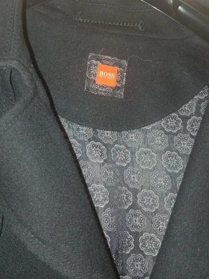 Svart trenchcoat från BOSS - Stilren svart trenchcoat från BOSS med dubbelknäppning och knytskärp i midjan. Jackan har klassisk krage och dekorativa knappar vid ärmsluten. Insidan är fodrad med ett blommigt mönster i grått. Perfekt för dig som vill ha en tidlös och elegant look.