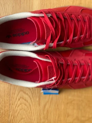 Röda Adidas sneakers i läder - Snygga röda Adidas sneakers med ovandel i läder och mockadetaljer. Skorna har vita innersulor och klassisk låg profil med matchande röda skosnören. Perfekta för dig som vill sticka ut med färg på fötterna.
