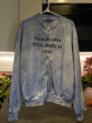 Ljusblå zip hoodie Acne Studios - Säljer en ljusblå zip hoodie från Acne Studios med texten 'Acne Studios STOCKHOLM 1996' på bröstet. Hoodien har dragkedja framtill, ribbade muddar och en avslappnad passform. Perfekt för dig som gillar streetwear och skandinavisk design.