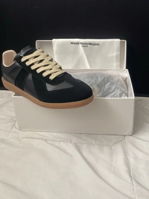 Maison Margiela skor  - Maison Margiela sneakers i svart läder och mocka med beigea skosnören och klassisk gummisula. Snygg låg modell med stilren design och vita detaljer på insidan. Perfekta för dig som gillar minimalistisk och trendig stil. De är för små på mig vilket är andledningen till attt jag säljer de.  Skorna är köpta på vinted och man får med förpackning. Ställ gärna eventuella frågor eller prisförslag.