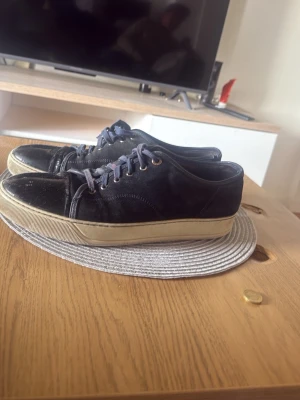 Lanvins  - Säljer ett par svarta sneakers med lågt skaft och vit, tjock sula. Skorna har svarta och mörkblå skosnören samt metallfärgade öljetter. Ovandelen verkar vara i textil och syntetmaterial, med en klassisk och enkel design.