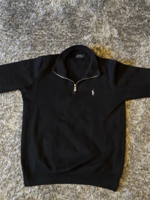 Svart halvzip tröja från Polo Ralph Lauren - Klassisk svart tröja från Polo Ralph Lauren med halvzip och hög krage. Tröjan har det ikoniska broderade logot på bröstet och ribbade muddar vid ärmslut och nederkant. Perfekt för en stilren och avslappnad look.
