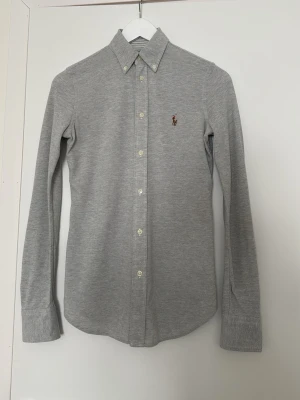 Grå skjorta från Ralph Lauren - Klassisk grå skjorta från Ralph Lauren med knappslå och button-down krage. Skjortan har långa ärmar och den ikoniska broderade loggan på bröstet. Tillverkad i mjuk bomull för en bekväm känsla och stilren look.