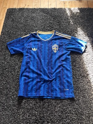 Sverige fotbollströja VM 2026 - Säljer en Sverige VM 2026 fotbollströja från Adidas med Sveriges landslagsemblem på bröstet. Tröjan har guldfärgade detaljer, klassiska Adidas-ränder på axlarna och ett unikt mönster i olika blå nyanser. Perfekt för match eller träning.
