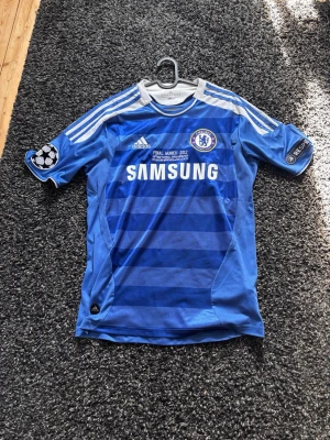 Chelsea FC 2011/2012 fotbollströja ”Final Munich 2012”  - Säljer en riktigt fet Chelsea FC matchtröja från Adidas i blått med vita detaljer och tryck. Tröjan har Champions League-finalen 2012-tryck, klubbmärke, sponsorlogga och UEFA Respect-märke på ärmen. Tillverkad i lätt och ventilerande material, perfekt för fotboll eller samling.