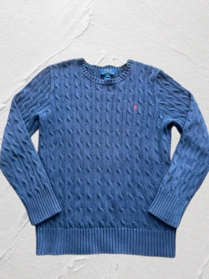 Blå kabelstickad tröja Polo Ralph Lauren - Kabelstickad blå tröja från Polo Ralph Lauren med rund halsringning och ribbade muddar. Tröjan har en liten rosa broderad logga på bröstet och är tillverkad i mjukt material. Perfekt för dig som gillar klassisk och stilren look.