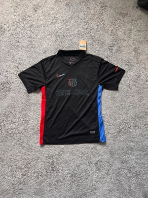 Svart FC Barcelona tröja Nike Travis Scott - Snygg limited edition Travis Scott svart FC Barcelona tröja från Nike med klubbmärke på bröstet och färgdetaljer i rött och blått på sidorna. Tröjan har korta ärmar, V-ringning och tryck på framsidan. Perfekt för fotboll eller träning. 