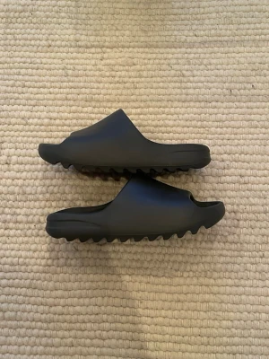 Yeezy Slides Onyx - Ett par Yeezy Slides Onyx i bra skick. Dom är använda och det finns därför vissa defekter, men för det mesta så måste man kolla noga i starkt ljus för att se dessa. Storlek : 44.5. Det rekommenderas att gå upp i storlek. Dom är 27.5 cm i längd där foten sitter.