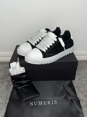 Svarta och vita sneakers från Numeris - Stilrena sneakers från Numeris i svart mocka med vita detaljer, tjock vit sula och vita skosnören. Skon har en låg profil och loggan är tryckt på hälen. Medföljer extra skosnören och dustbag. Perfekta för dig som gillar minimalistisk och modern stil.
