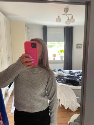 Grå stickad tröja med pärldetaljer - Snygg grå stickad tröja med pärldetaljer runt halsringningen. Tröjan har lång ärm och är något croppad i modellen. Perfekt för dig som vill ha en enkel men ändå lite extra stickad tröja.