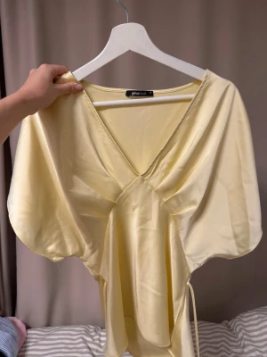 Ljusgul omlottopp i satin - Supersöt ljusgul omlottopp i satin med v-ringning och korta, vida ärmar. Toppen har knyt i sidan för en snygg passform och ett mjukt, glansigt material som känns lyxigt. Perfekt för dig som vill ha en feminin och trendig look.