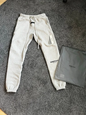Fear of God Essentials Sweatpants - These are grey Fear of God Essentials sweatpants, size XS. The pants feature the signature 'ESSENTIALS FEAR OF GOD' branding on the left thigh and a rubberized Essentials label on the front waistband. The sweatpants come with their original packaging, as shown in the first image.