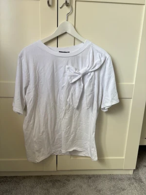 Vit t-shirt med rosett från New Collection - Säljer en vit t-shirt från New Collection med en stor rosett på axeln som ger en unik touch. T-shirten har korta ärmar och är gjord i mjuk bomull. Perfekt för dig som vill ha något enkelt men ändå speciellt i garderoben.