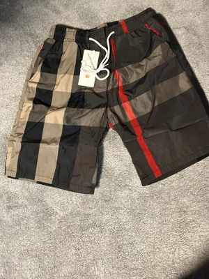 Rutiga badbyxor med röd detalj - Snygga badbyxor med klassiskt rutigt mönster i svart, beige och grått samt en tydlig röd rand framtill. De är sköna och de kan användas som vanliga shorts även kan diskutera pris vid snabb affär m/s 