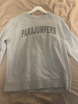 Ljusblå sweatshirt från Parajumpers - Ljusblå sweatshirt från Parajumpers med stor logga i grått på bröstet. Tröjan har rund halsringning, ribbade muddar vid ärmslut och nederkant samt en liten patch på ena ärmen. Perfekt för en avslappnad och stilren look.