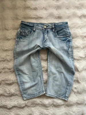 Ljusblå Capri jeans  - Säljer ett par lågmidjade ljusblå capri jeans med rak passform och klassisk femficksdesign. Jeansen har slitna detaljer och är tillverkade i denim med en skön, avslappnad look. Perfekta för dig som gillar en casual stil.