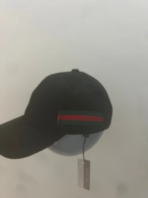 Svart keps G med röd-grön rand - Snygg svart keps från G med klassisk röd och grön rand på sidan. Kepsen har böjd skärm och justerbar rem bak. Tillverkad i ett slitstarkt material och har en stilren look som passar till många outfits.