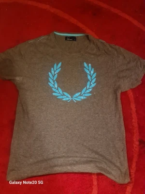 Grå t-shirt från Fred Perry - Säljer en grå t-shirt från Fred Perry med den klassiska blå lagerkransen tryckt på bröstet. T-shirten har rund hals och korta ärmar. Perfekt för dig som gillar stilrena och sportiga plagg.