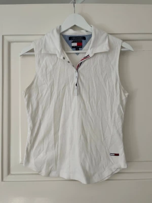 Vit ärmlös piké från Tommy Jeans - Säljer en vit ärmlös pikétröja från Tommy Jeans med klassisk krage och knappslå framtill. Tröjan har en liten logga nertill och snygga rödblå detaljer vid knapparna. Perfekt till jeans eller kjol för en sportig look. Passar XS/S