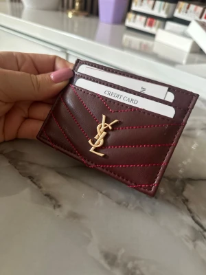 Vinröd kortfodral från Saint Laurent - Supersnyggt vinrött kortfodral från Saint Laurent i läder med guldigt YSL-logo framtill. Fodralet har flera kortfack och coola röda sömmar i chevronmönster. Perfekt för dig som vill ha stilren och lyxig accessoar i fickan.