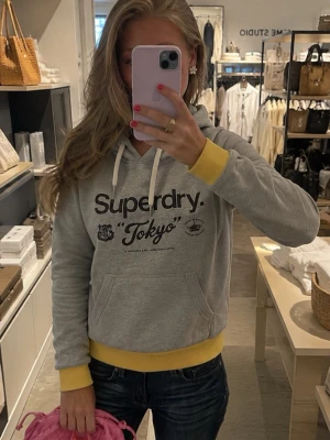 Grå Superdry hoodie med gul kant - Snygg grå hoodie från Superdry med svart text och tryck på bröstet. Hoodien har huva med vita snören, känguruficka och kontrasterande gula muddar och nederkant. Perfekt för en chill och avslappnad stil.