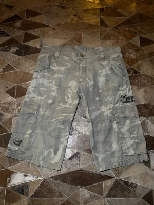 Grå camouflage cargo shorts - Säljer ett par gråa cargo shorts med camouflage-mönster. Shortsen har flera fickor på sidorna och bak, samt knapp och dragkedja framtill. Perfekta för en avslappnad och cool stil.